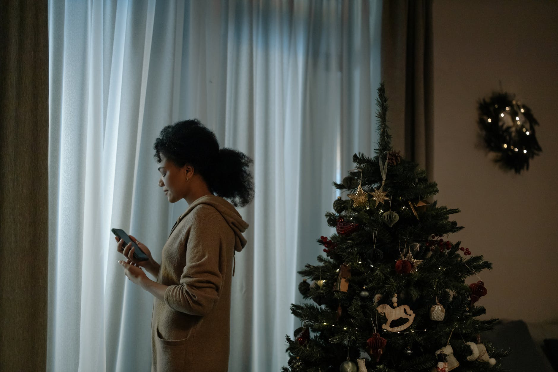 Apart at Christmas - A woman standing alone looking down sadly at a cell phone. In back of her is a Christmas tree.