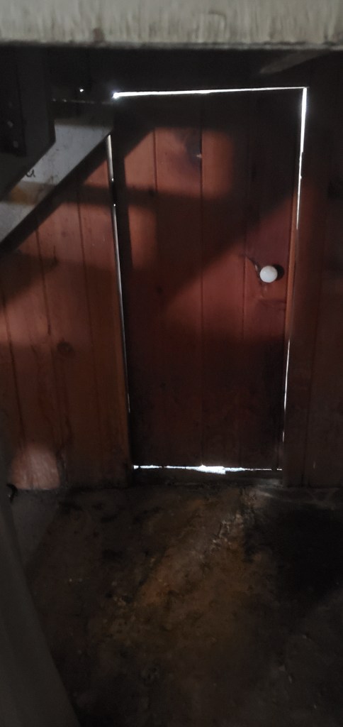 Photo shows a small wodden door with an old-fashioned knob. It is located under a set of wooden stairs and has light shining through from the other side.