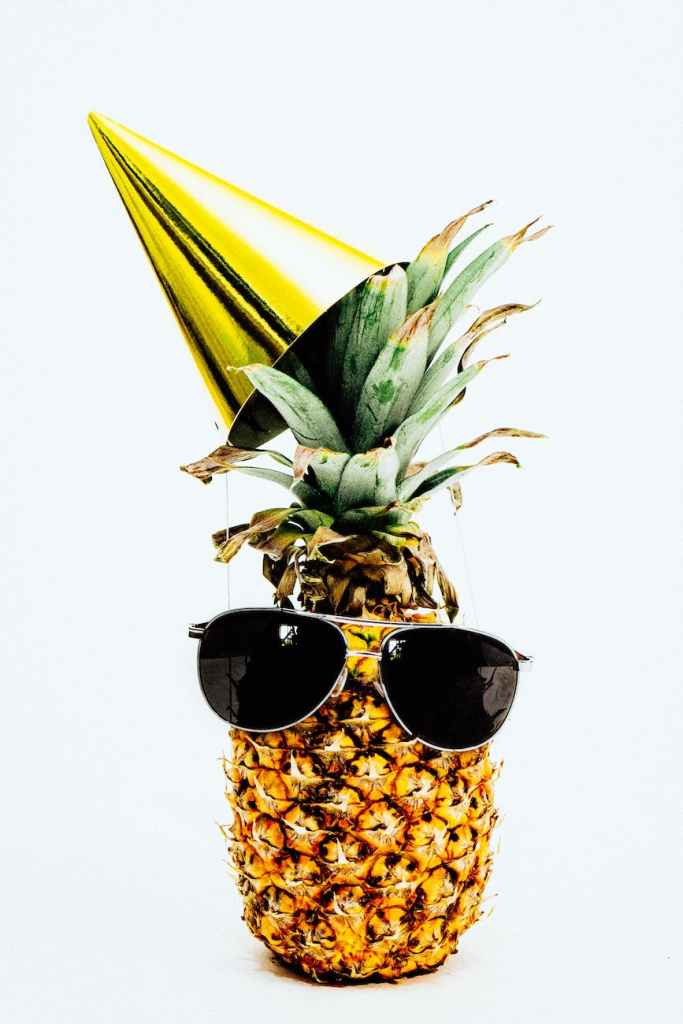 Image shows a pineapple with a gold party hat and aviator-type sunglasses.