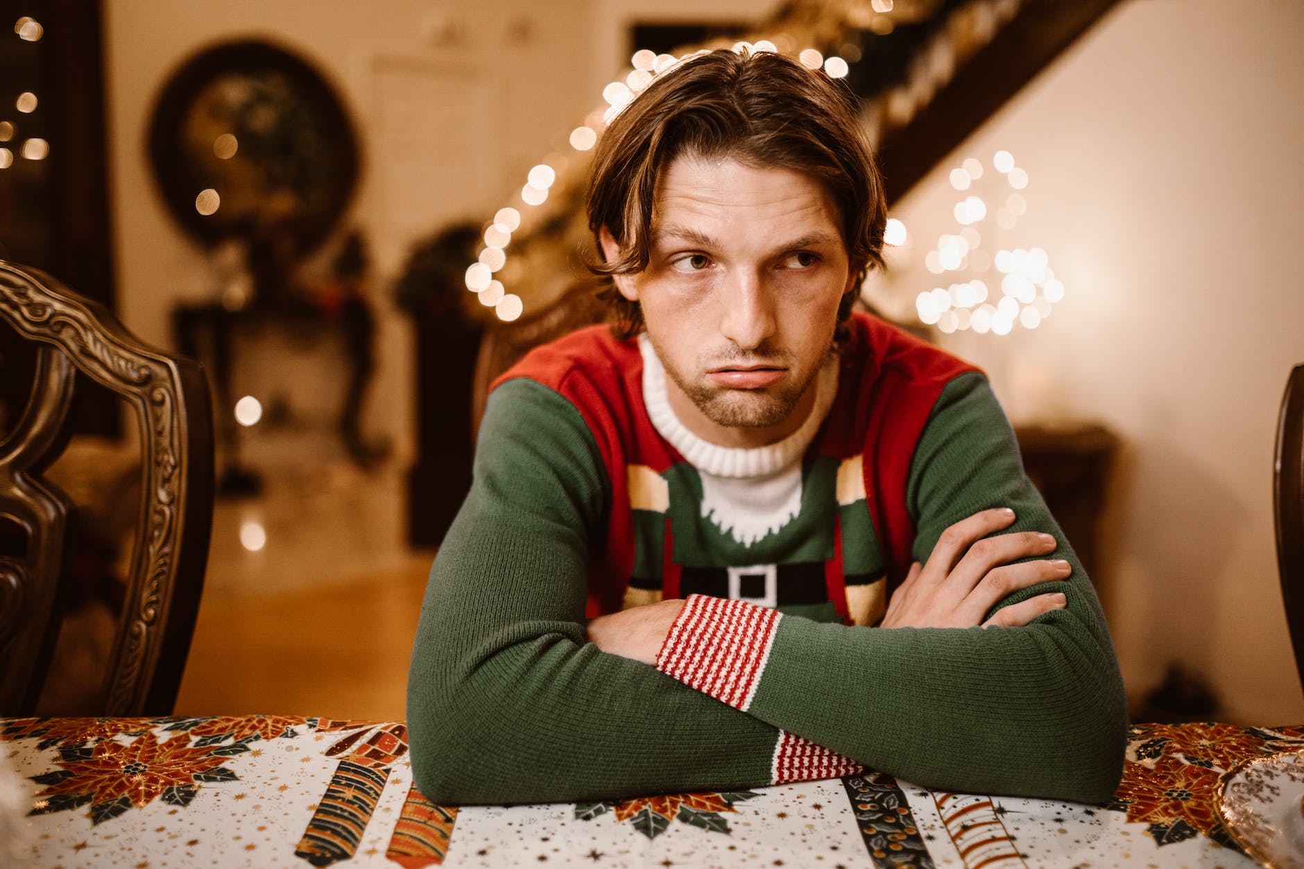Image shows a man in a green, red and white sweater with his arms folded on a table with a "holiday" table cloth. He appears depressed and in the blurred background are holiday lights and decorations.
