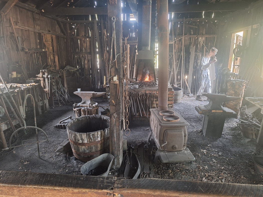 The inside of the blacksmith’s shop with various equipment all around the room and hanging from the ceiling, a young man in one corner of the room, and a couple of wood stoves.